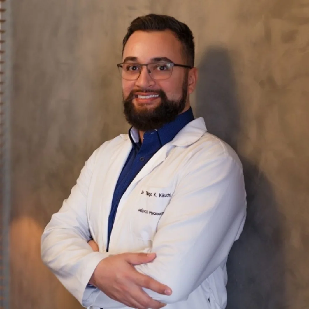 Dr. Thiago Koiti Kikuchi de jaleco, braços cruzados, diante da parede de cimento queimado